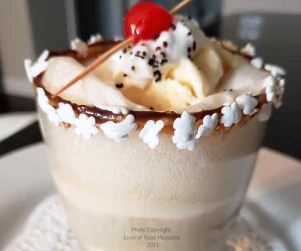 Ice Cream floating in a glass with whipped cream, a bright red cherry and little candies rim the glass over some chocolate syrup